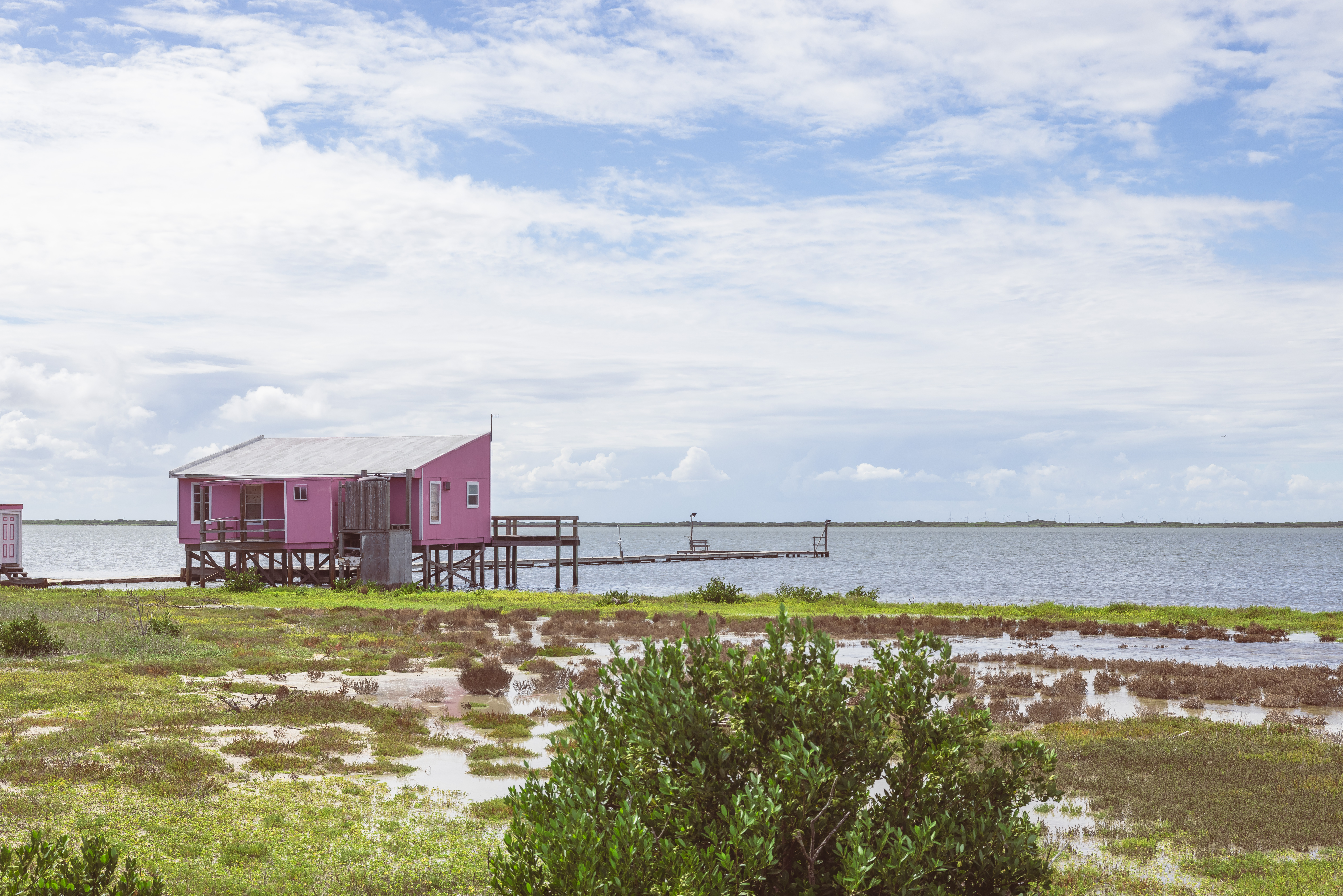 The image shows the back of a pink house sitting on top of stilts holding it above water on the edge of the Laguna Madre. The house faces out towards the water.